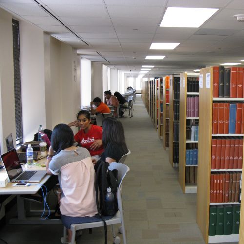 Students sit at tables in the library, to the right are bookshelves.