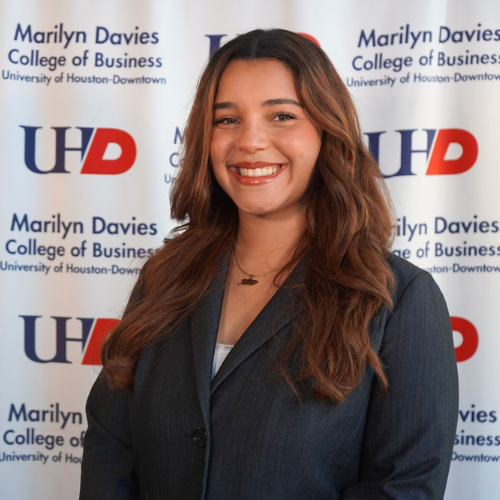 Senior and Ted Bauer Scholar Cameliss Rivera Gautier smiles in front of a backdrop with the University of Houston-Downtown and Marilyn Davies College of Business logos. 