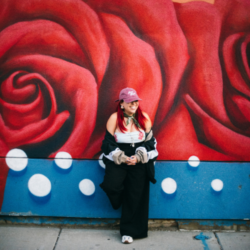 Magge Nunez a University of Houston-Downtown alumna smiles with a mural of a rose behind them. Nunez is a DJ in Houston, Texas.